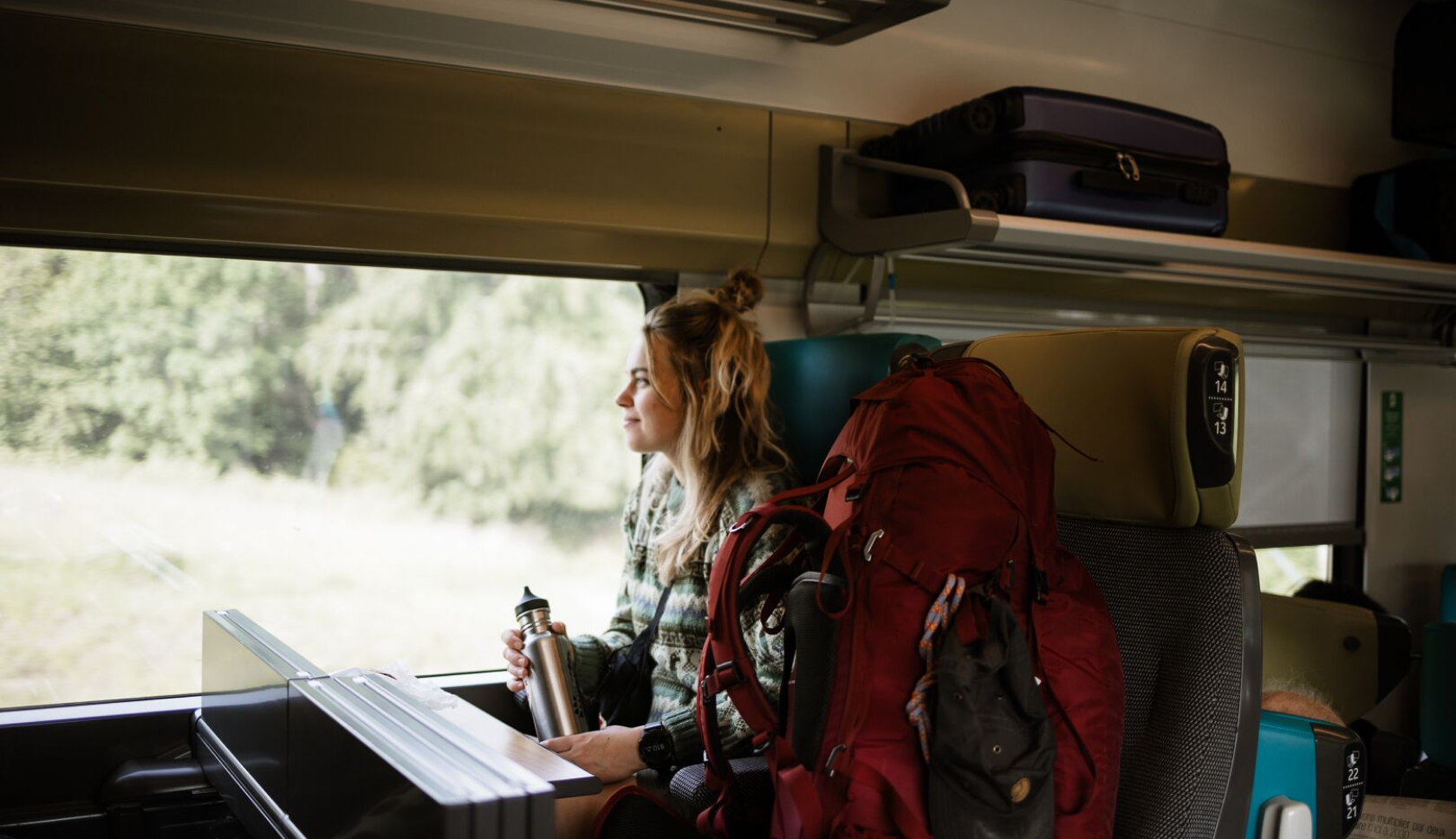 Een vrouw zit in een trein met een grote, rode backpack naast zich