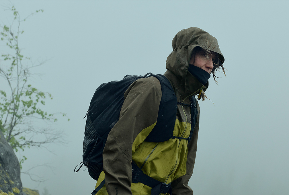 vrouw in groene jas met capuchon op en een zwarte rugzak op haar rug