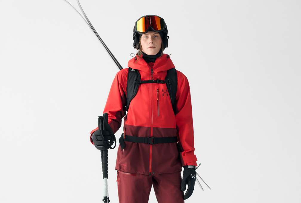 vrouw in wintersportoutfit staat met skistokken in de hand tegen een witte achtergrond