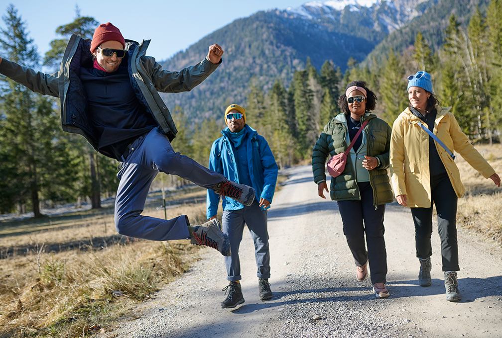 vier mensen wandelen met bergen op de achtergrond, voorste man springt vrolijk in de lucht